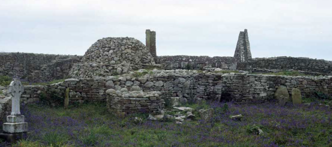Ancient stone ruins with circular and rectangular structures, an old sweathouse, gravestones, and wild purple flowers growing among the grass in the foreground under a cloudy sky.