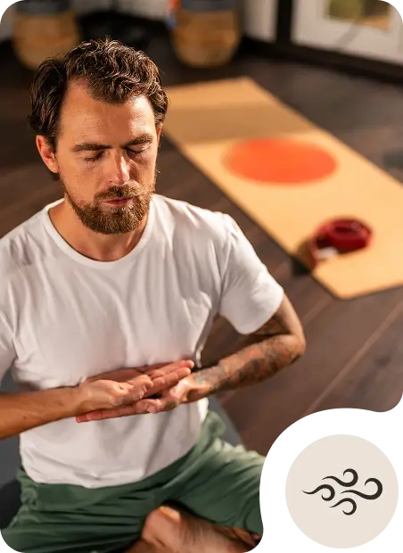 A bearded man in a white t-shirt and green pants sits cross-legged on a yoga mat indoors, meditating with eyes closed. In the background is another mat and yoga props. An air element symbol is overlaid on the image.