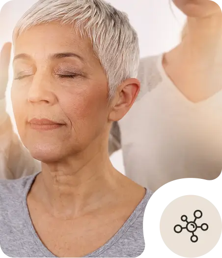 A serene older woman with short gray hair sits with her eyes closed while another person stands behind her, appearing to perform a healing or relaxation technique. An abstract icon is displayed in the bottom corner.