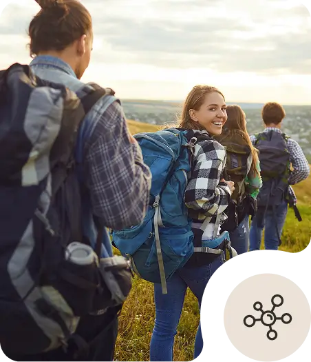 A group of four hikers with backpacks walk through a grassy field; one woman in a plaid shirt turns and smiles at the camera. The scene has warm lighting, and a circular network icon is overlaid in the corner.