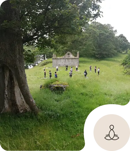 A group of people stands in a circle on grass near old stone ruins and trees, practicing yoga or meditation outdoors in a lush, green landscape. An icon of a meditating figure appears in the lower right corner.