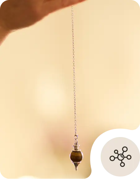 A hand holds a chain with a pointed pendulum suspended in the air against a soft, blurred beige background.