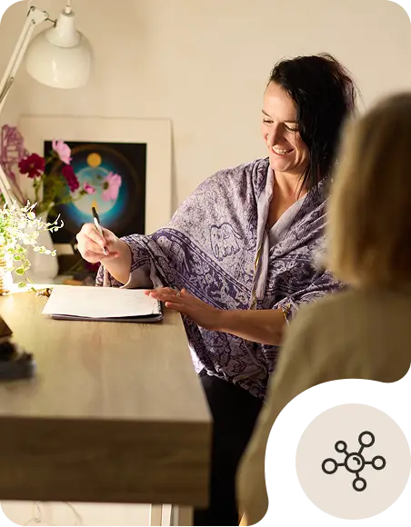 A woman wrapped in a purple shawl smiles while writing in a notebook at a desk, facing another person. A lamp, flowers, and colorful artwork are in the background.