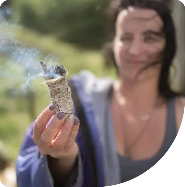 A woman with dark hair holds a smoking bundle of sage outdoors, with blurred greenery in the background. She wears a blue robe and a grey top, and her nails are painted grey.