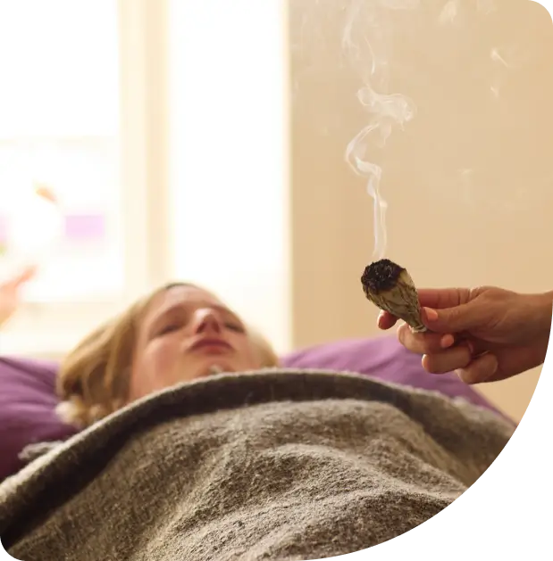 A person lies on a bed covered with a blanket while another hand holds a smoldering bundle of sage nearby, with smoke wafting in the air, suggesting a cleansing or relaxation ritual.
