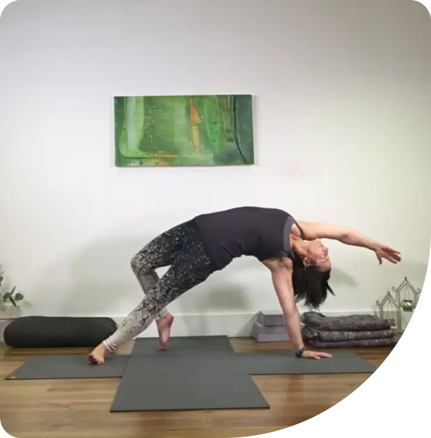 A woman in yoga attire performs a side plank pose on a mat indoors, with one hand and foot on the ground, her other arm extended overhead. A green painting, yoga props, and a plant are visible in the background.