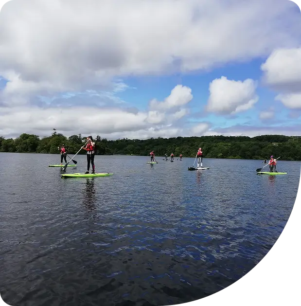 A group of people wearing red life jackets stand on paddleboards on a calm lake, surrounded by trees under a partly cloudy sky.