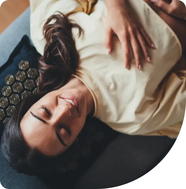 A woman with long dark hair lies on her back on a couch, smiling with her eyes closed and one hand resting on her stomach, appearing relaxed and content.