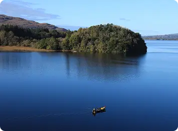 A small boat with two people floats on a calm blue lake surrounded by lush green trees and hills under a clear sky.