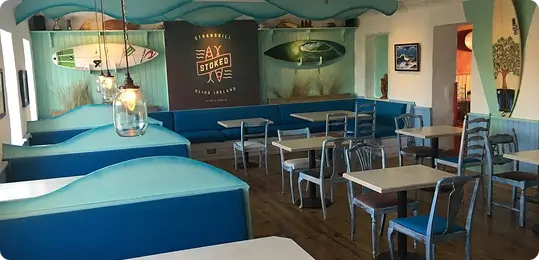 A brightly lit beach-themed cafe with blue booths and chairs, surfboards and ocean decor on the walls, and a sign reading “Stoked” above the seating area.