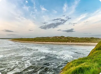A scenic coastal view showing waves crashing onto a sandy beach, with grassy dunes in the background and a partly cloudy sky overhead.