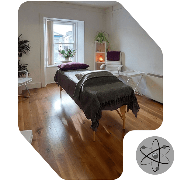 A cozy treatment room with a massage table covered in a dark blanket, plants by a window, a white chair, and a small shelf with a glowing lamp. A science-themed atom symbol is in the bottom right corner.