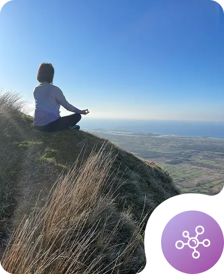 A person sits cross-legged on a grassy hillside, meditating and focusing on wellbeing while facing a scenic view of fields and the ocean under a clear blue sky. A circular digital icon with network nodes appears in the bottom right corner.
