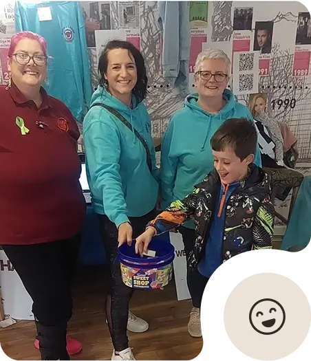 Four smiling adults and one smiling child stand indoors. The child is reaching into a blue bucket labeled Sweet Shop. The adults wear casual clothes, including hoodies and a polo shirt. A display and posters are visible in the background.