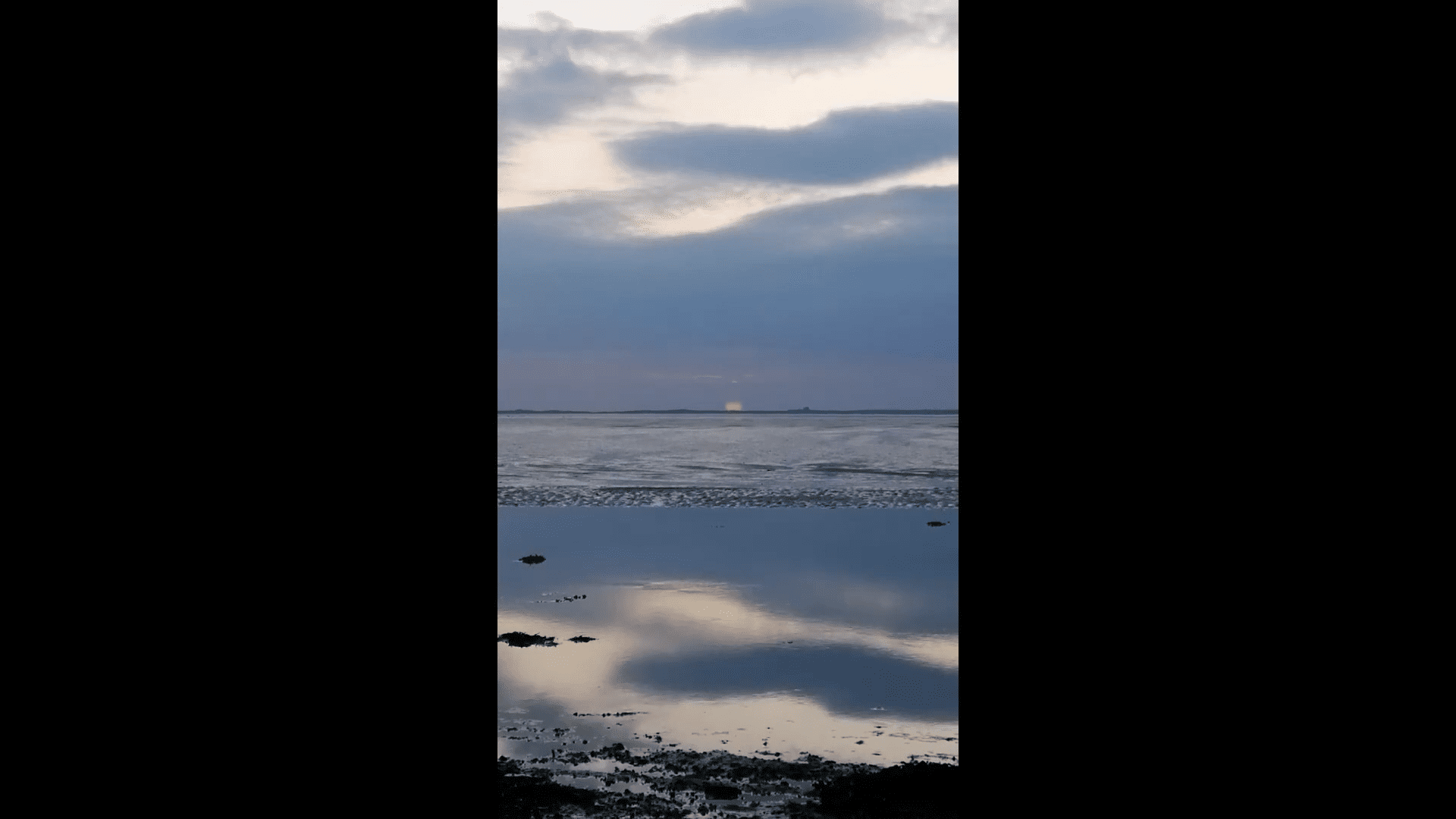 A calm seascape at dusk with clouds in the sky, a faint distant light on the horizon, and the sky reflected in the still water below.