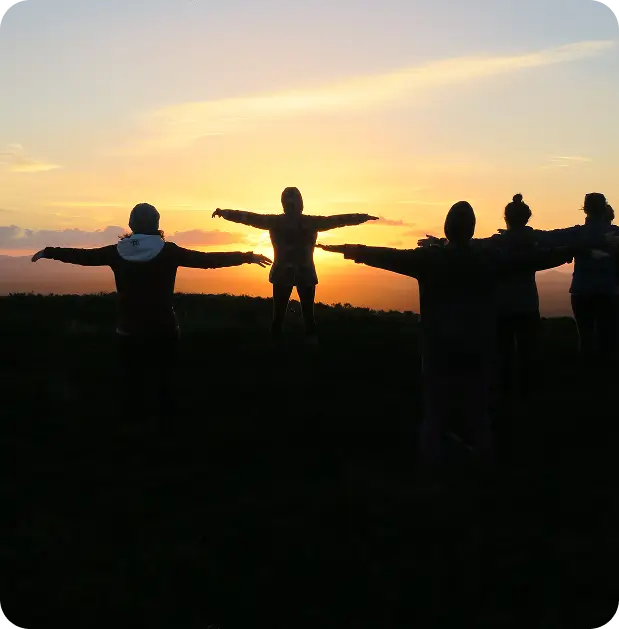 Five people stand outdoors with arms outstretched, silhouetted against a sunset sky with orange and yellow hues. The group appears to be enjoying the scenic view together.