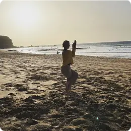 A person practices yoga on a sandy beach at sunset, standing on one leg with arms raised. The ocean and a distant cliff are visible in the background.