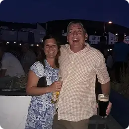 A woman and a man stand close together outdoors at night, both smiling. The woman holds a bottle, and the man holds a pint of dark beer. There are people and houses in the dark background.