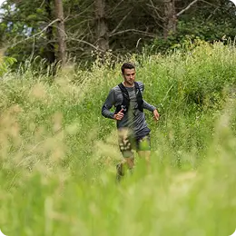 A man running through tall grass in a forested area, wearing athletic gear and holding a water bottle.