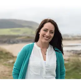 A woman with long brown hair, wearing a white blouse and teal cardigan, smiles while standing outdoors on a grassy landscape with hills and a beach in the background.