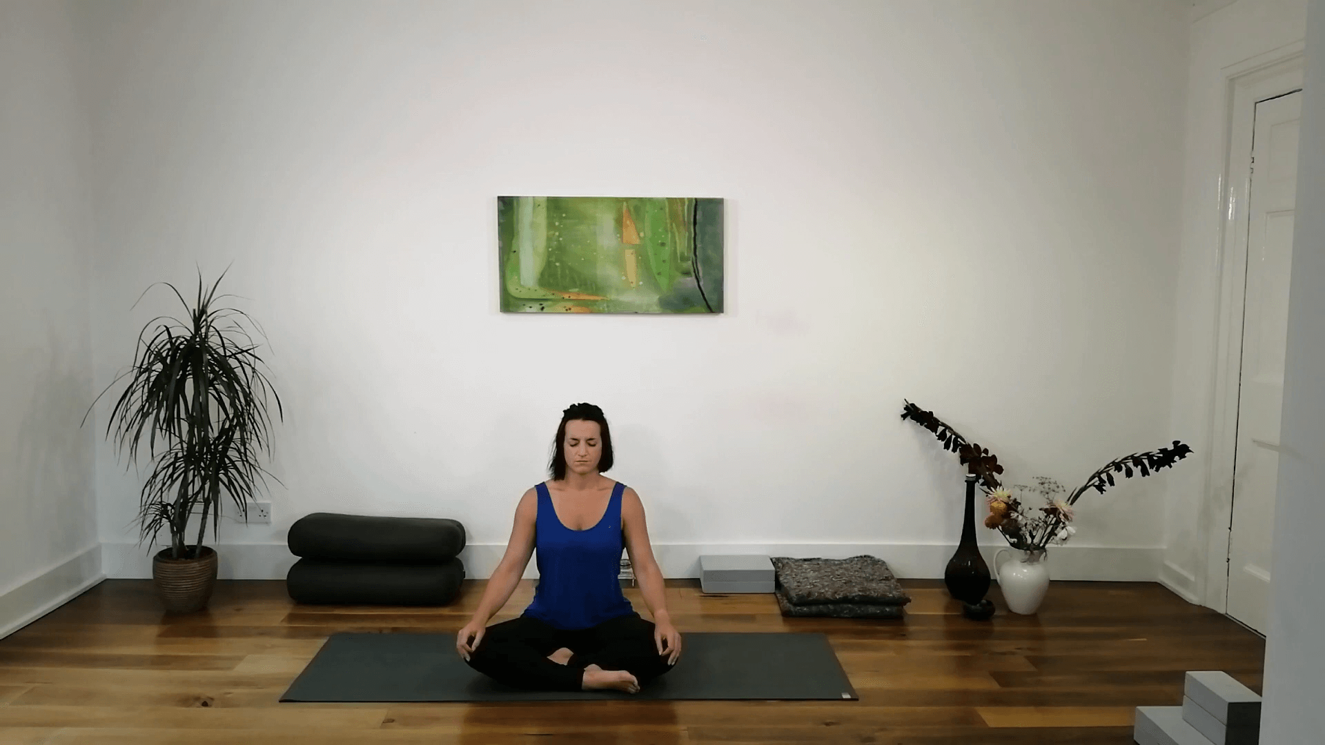 A woman sits cross-legged on a yoga mat in a minimalist room, meditating with eyes closed. Cushions, a potted plant, folded blankets, and decorative vases are arranged along the wall behind her.