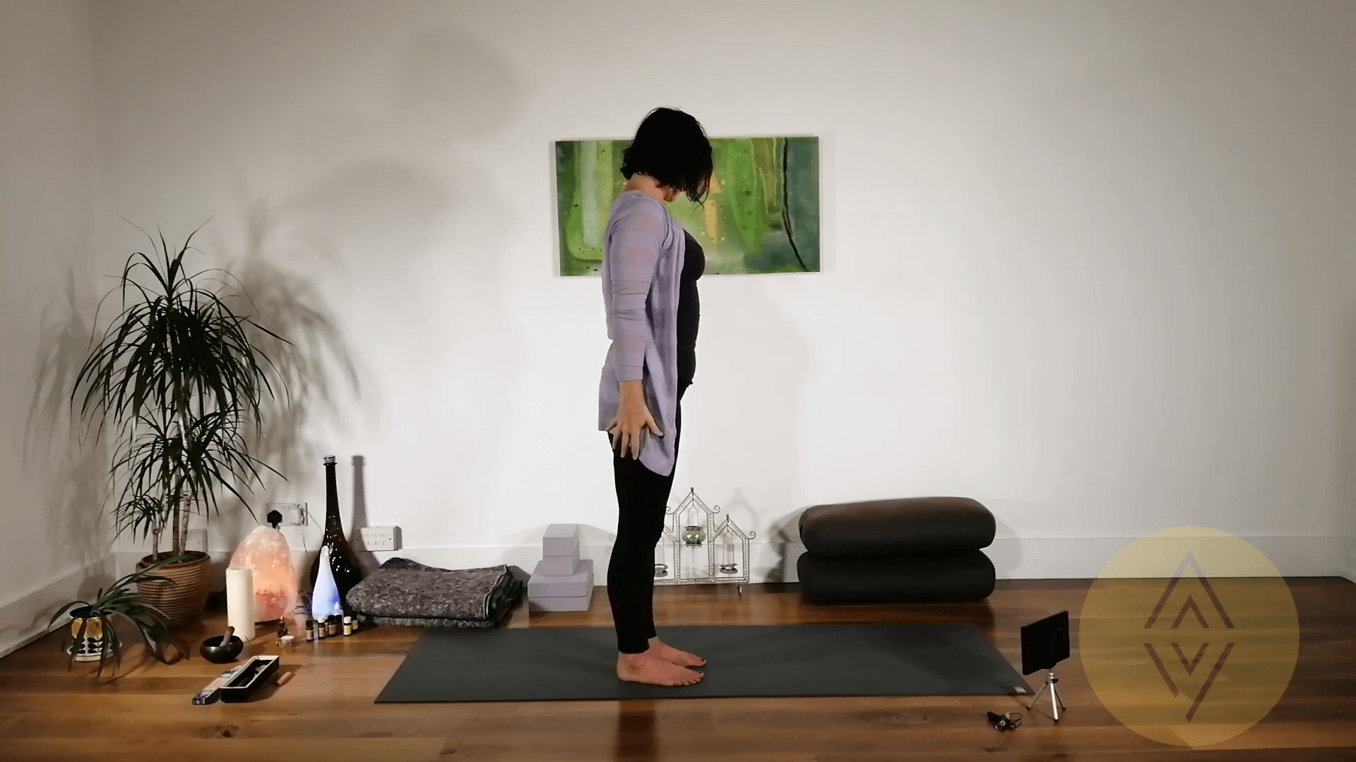 A woman stands upright on a yoga mat in a well-lit room, facing left. Yoga props, cushions, and a small plant are arranged neatly around her. A green painting hangs on the wall behind her.