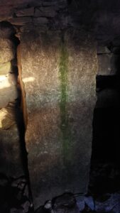 A large upright stone stands inside the chamber of a Carrowkeel passage tomb, illuminated by a narrow beam of sunlight entering from the left. The ancient stone surface shows natural weathering and a streak of green moss, while the surrounding dry-stone structure remains in shadow, highlighting the interplay of light and darkness within the tomb.