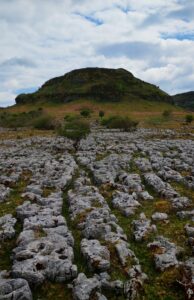 A limestone pavement stretches across the foreground beneath a rounded green hill. Weathered grey rocks, scattered shrubs, and patches of grass create a striking natural pattern under a dramatic sky filled with clouds and glimpses of blue.
